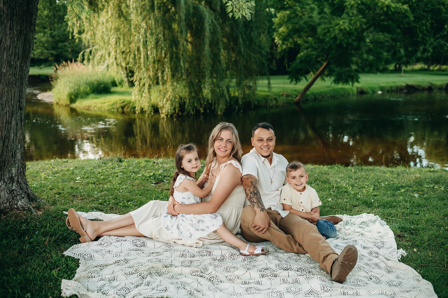 Family of four sitting on blanket by pond under willow tree in lush green park for outdoor portrait.