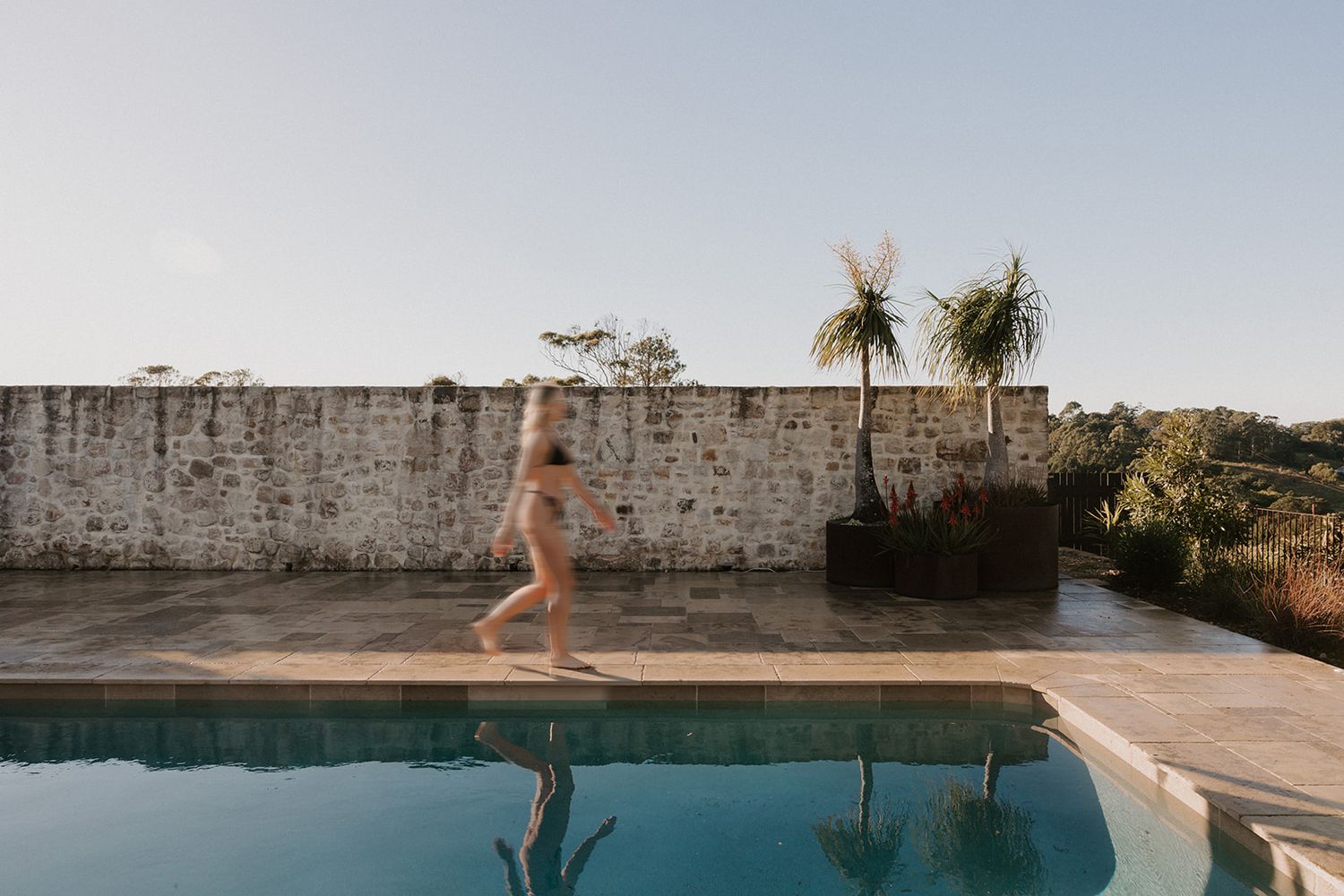 A person in a bikini running along a serene outdoor swimming pool beside a rustic stone wall with tropical plants.