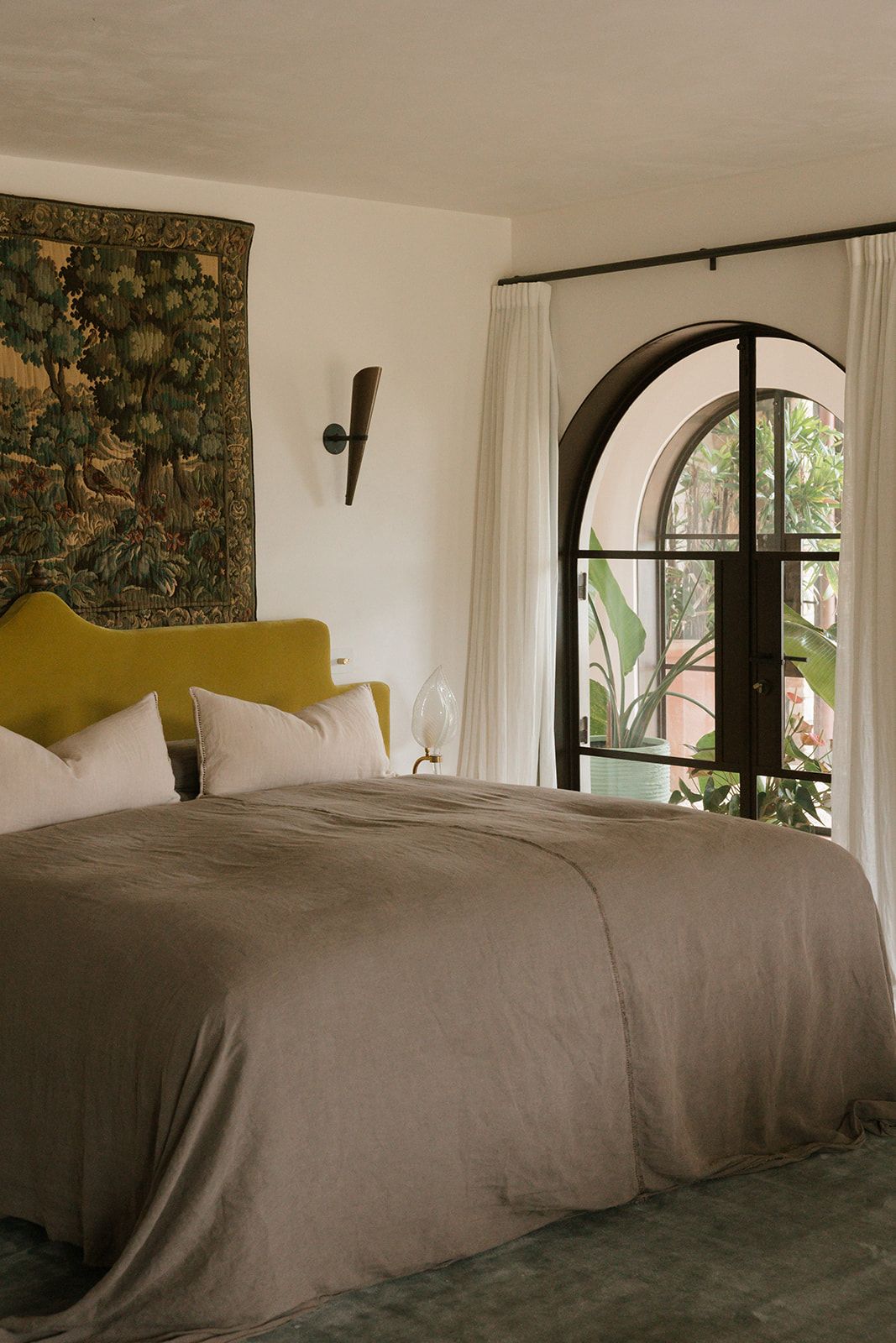 Elegant bedroom with olive velvet headboard, taupe linen bedding, arched window, and vintage tapestry wall art.