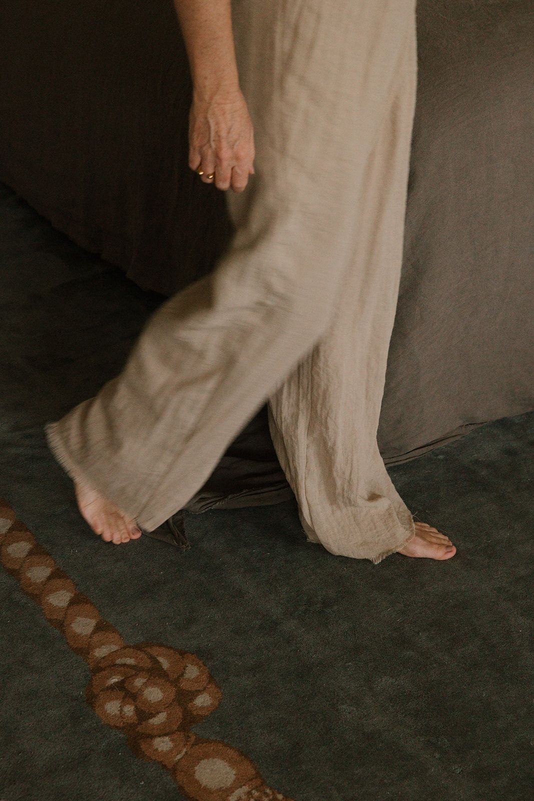 Person wearing wide-leg linen trousers walking barefoot on dark green carpet near decorative circular pattern.