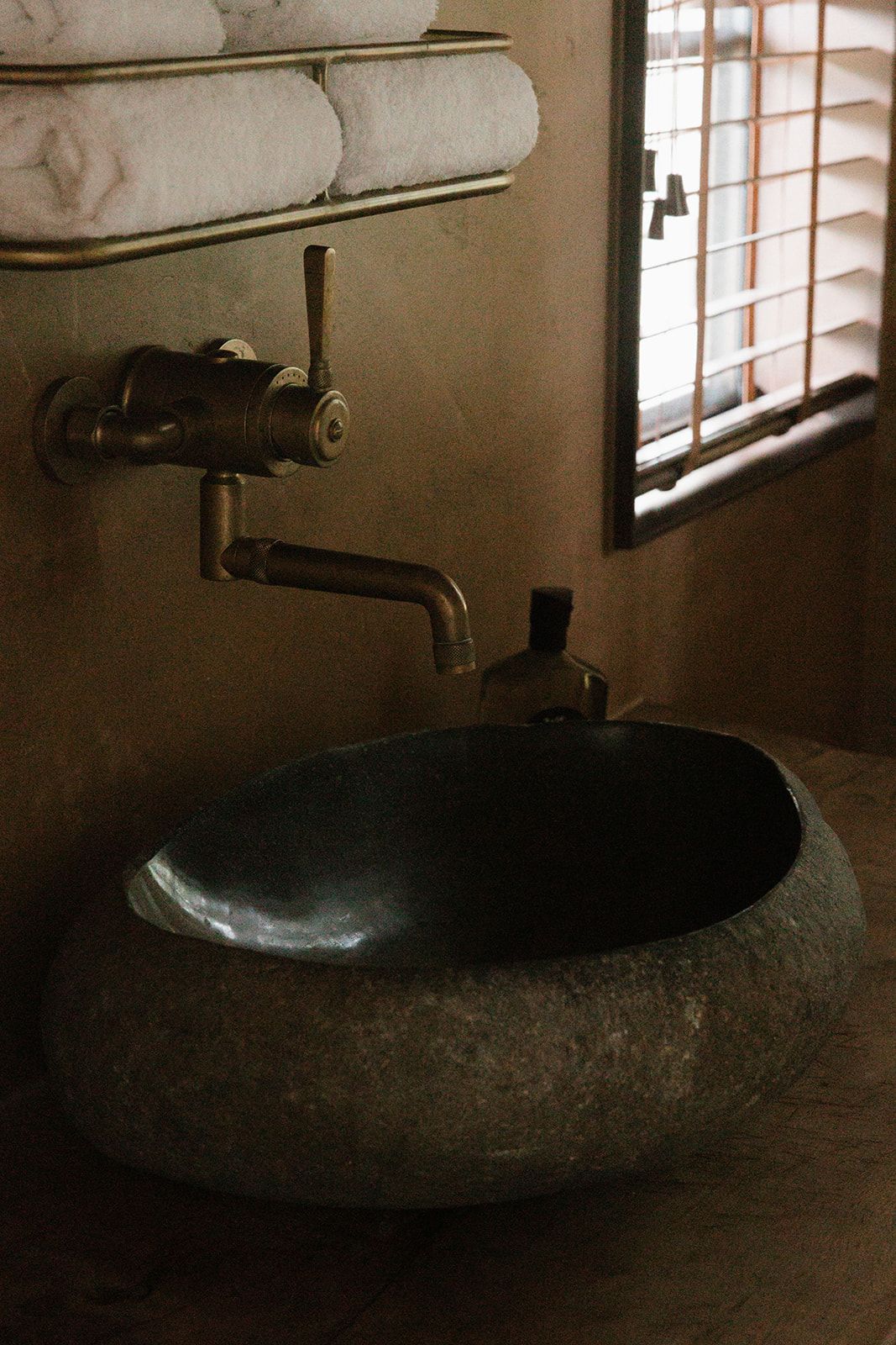 Rustic stone vessel sink with brass wall-mounted faucet, folded white towels on rack, near a wooden window.