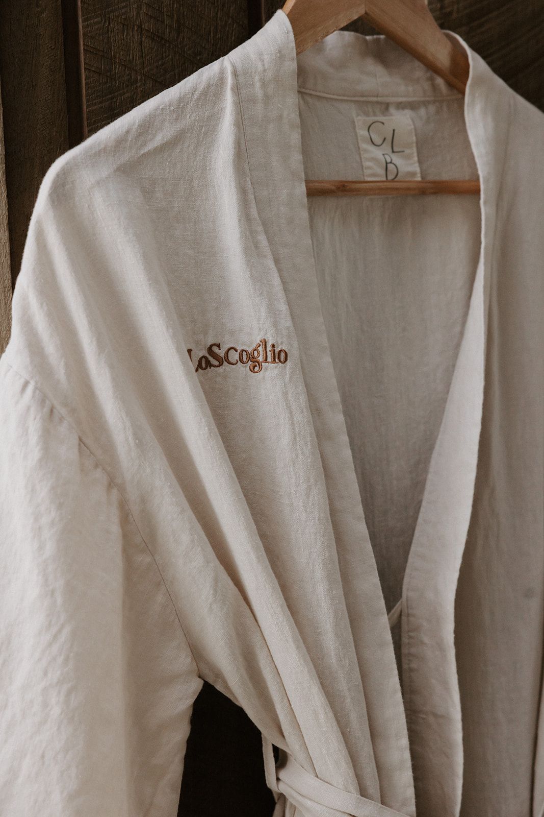 White linen Lo Scoglio branded hotel bathrobe hanging on a wooden hanger.