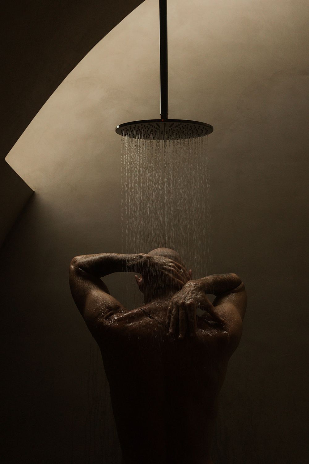 Person standing under a round rainfall shower head in a dimly lit, modern bathroom with dramatic lighting.