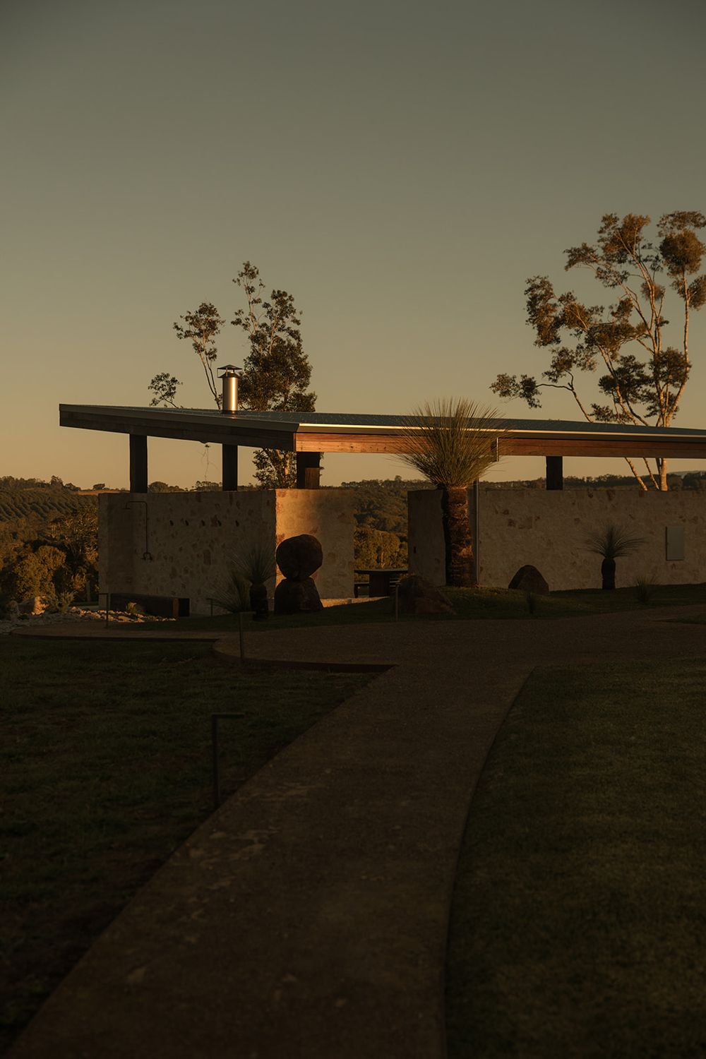 Modern stone building with flat roof at golden hour sunset, surrounded by eucalyptus trees and manicured lawn.