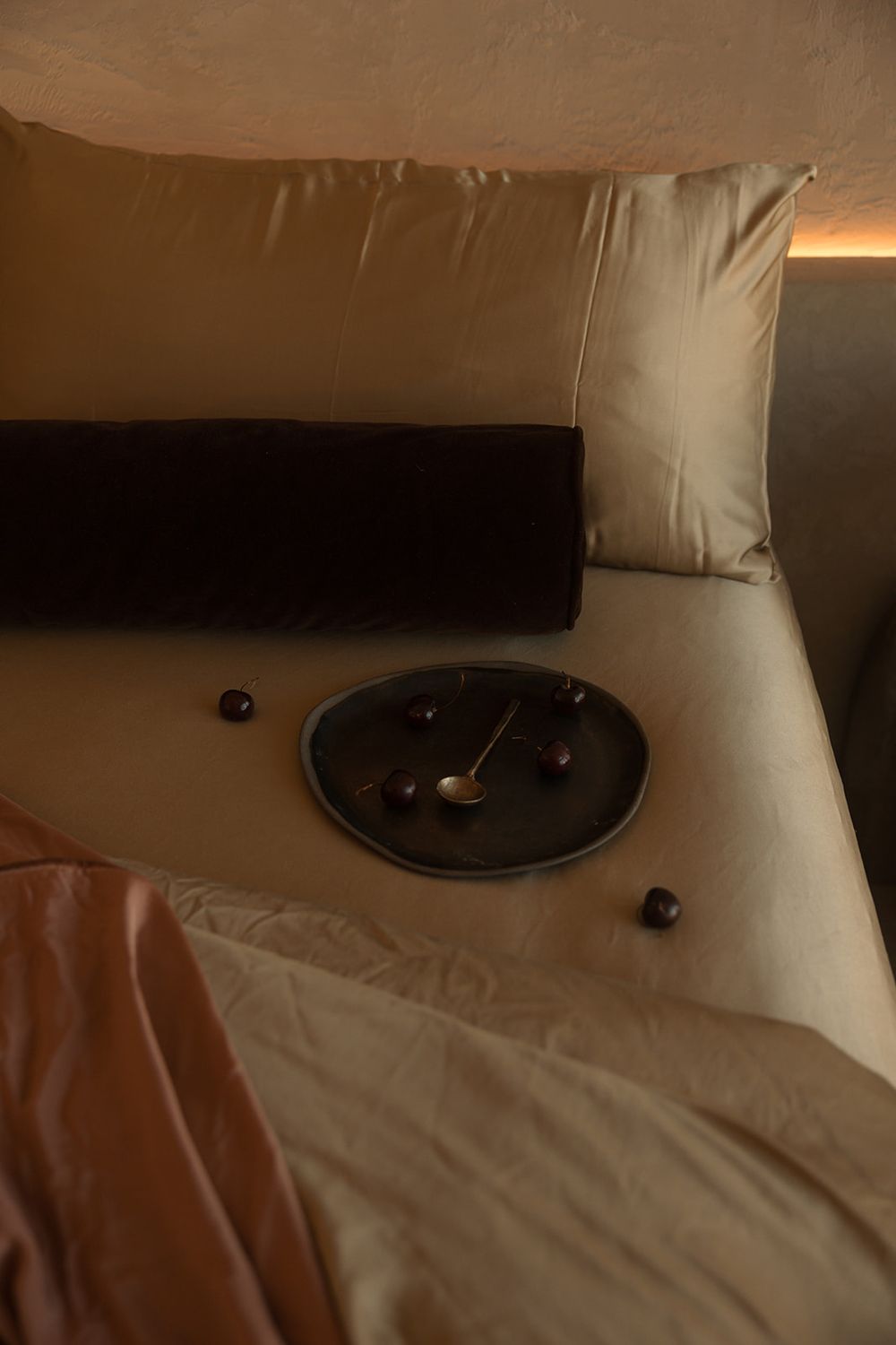 Luxurious bedroom with golden satin pillows, dark bolster, and elegant clock on beige bedding in warm ambient lighting.