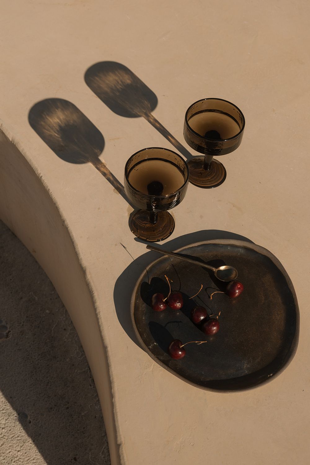 Two smoky brown coupe glasses and a dark plate with cherries cast dramatic shadows on a warm beige wall.