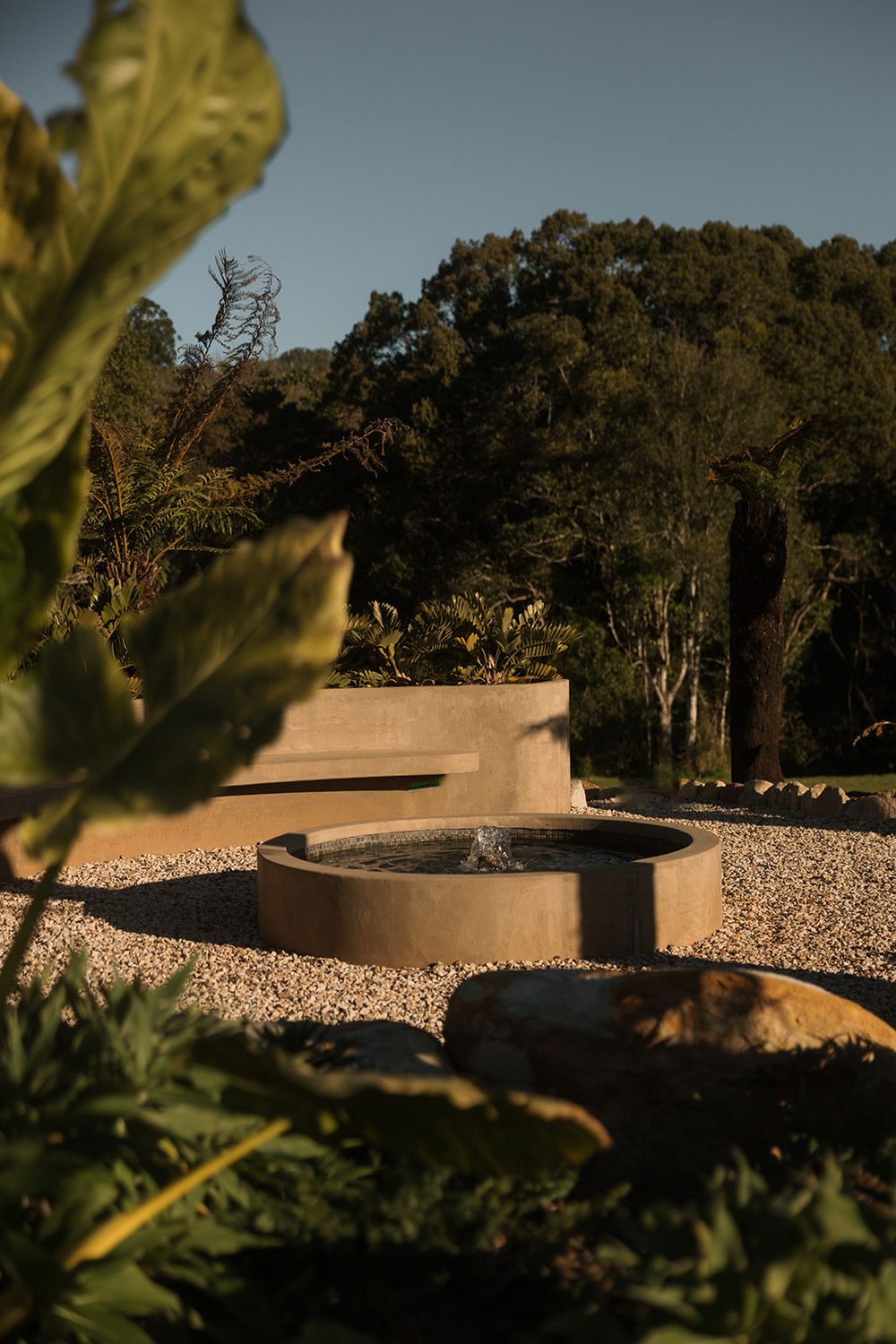 A serene garden fountain with circular concrete basin surrounded by gravel paths, lush greenery, and trees at golden hour.