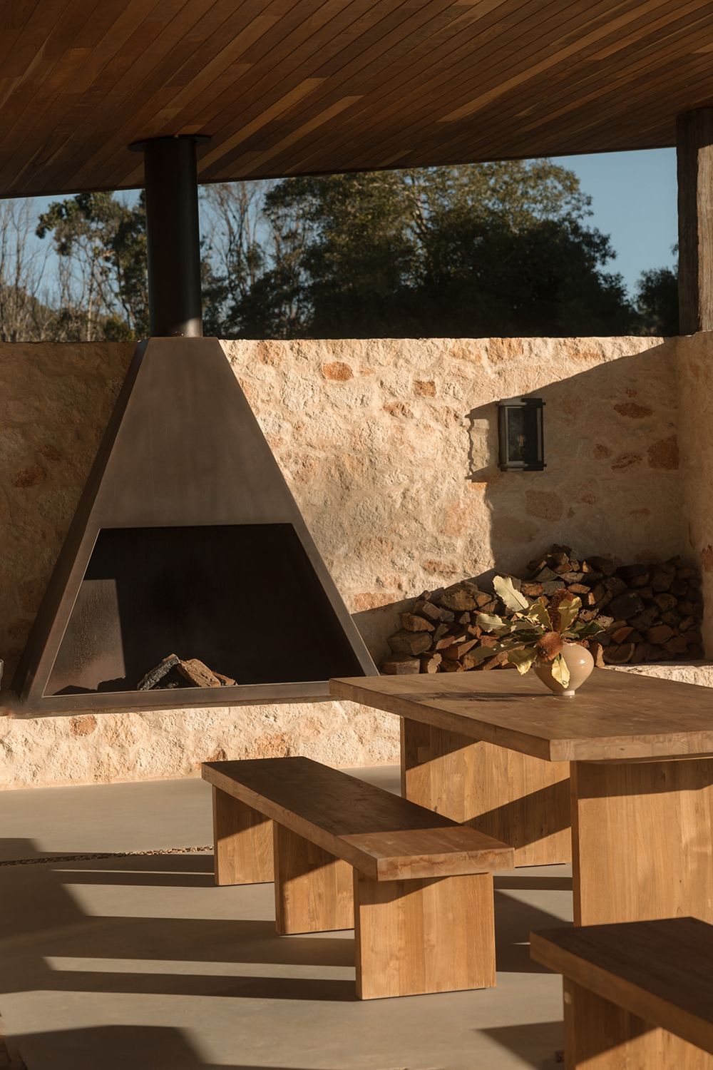 Rustic outdoor dining area with wooden table and benches, stone wall, firewood stack, and modern triangular fireplace under timber ceiling.