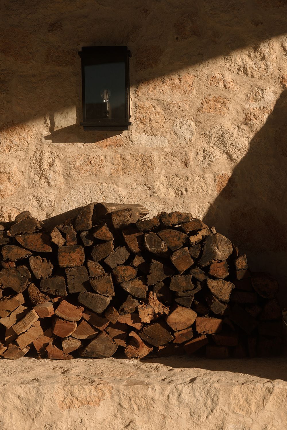 Rustic stone wall with stacked firewood logs in warm sunlight, featuring a small black wall lantern casting dramatic shadows.