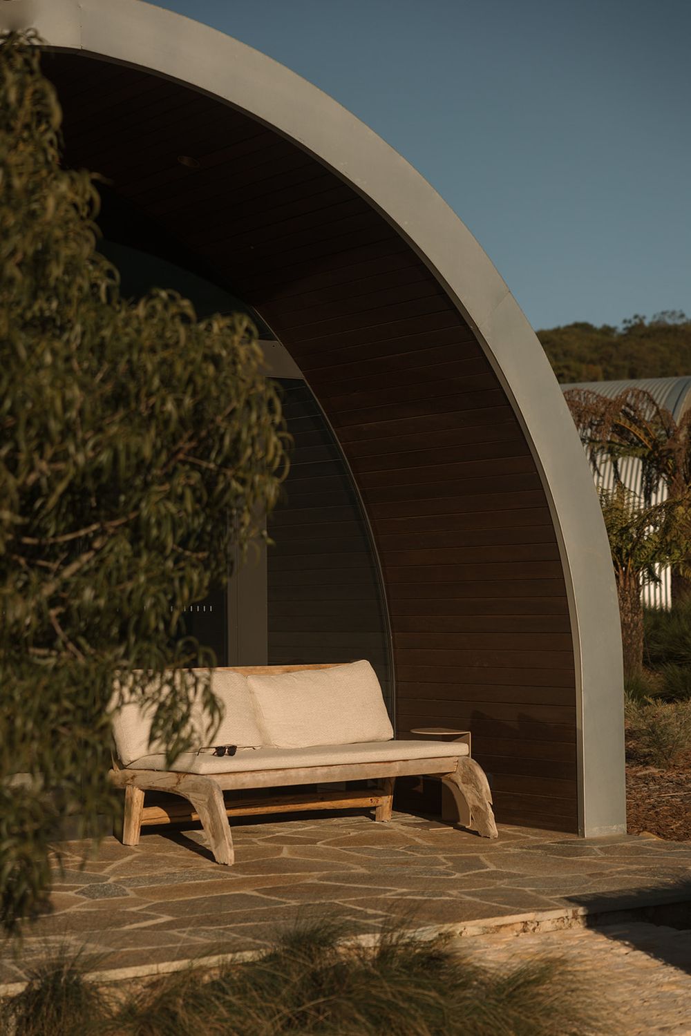 Rustic wooden bench with cushions under a curved concrete arch structure on a stone patio surrounded by native Australian plants.
