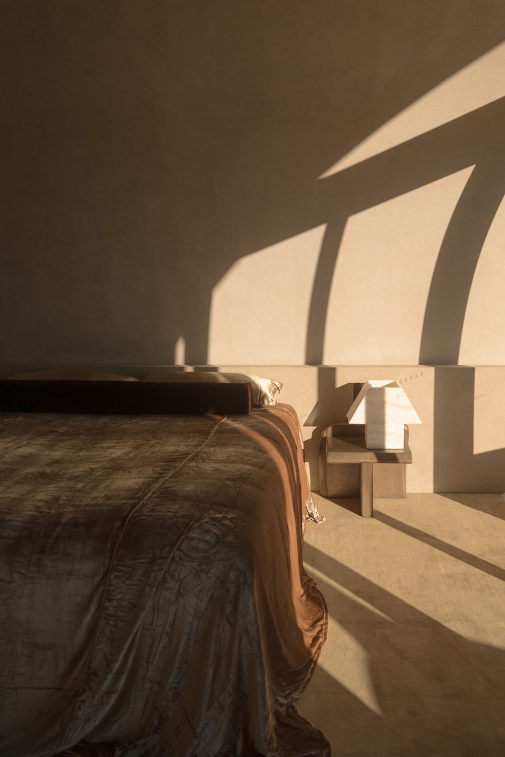 Warm sunlit bedroom with earth-tone bedding, concrete headboard, and small white lamp casting geometric shadows on beige walls.
