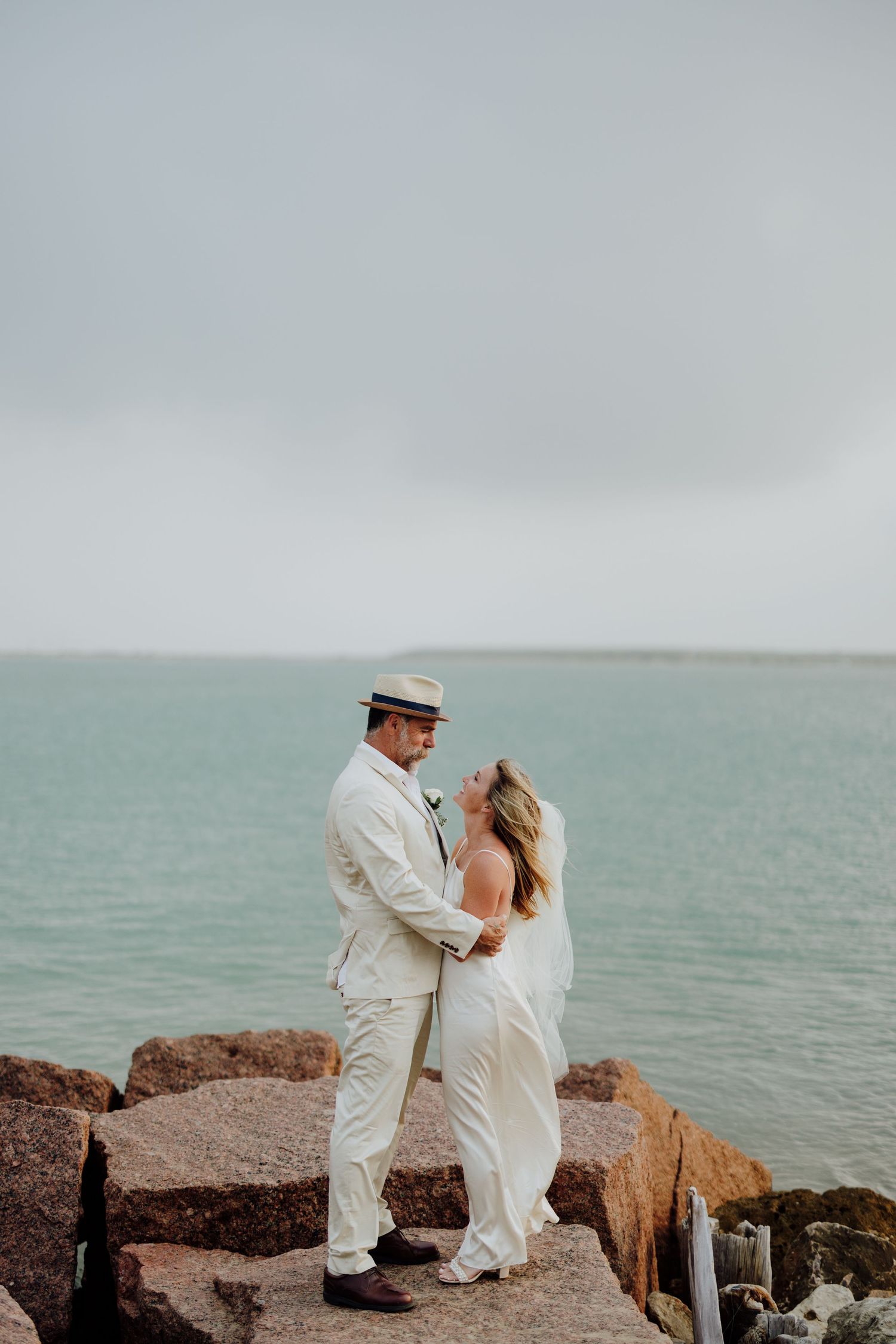 Couple in wedding attire embrace on rocky shoreline with turquoise ocean backdrop on cloudy day.