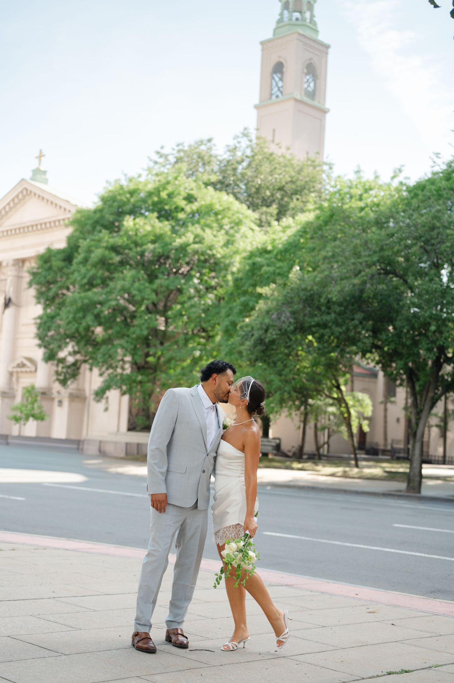 Newlywed couple sharing a kiss on a sunlit street with a historic church and lush green trees in the background in White Plains, NY.