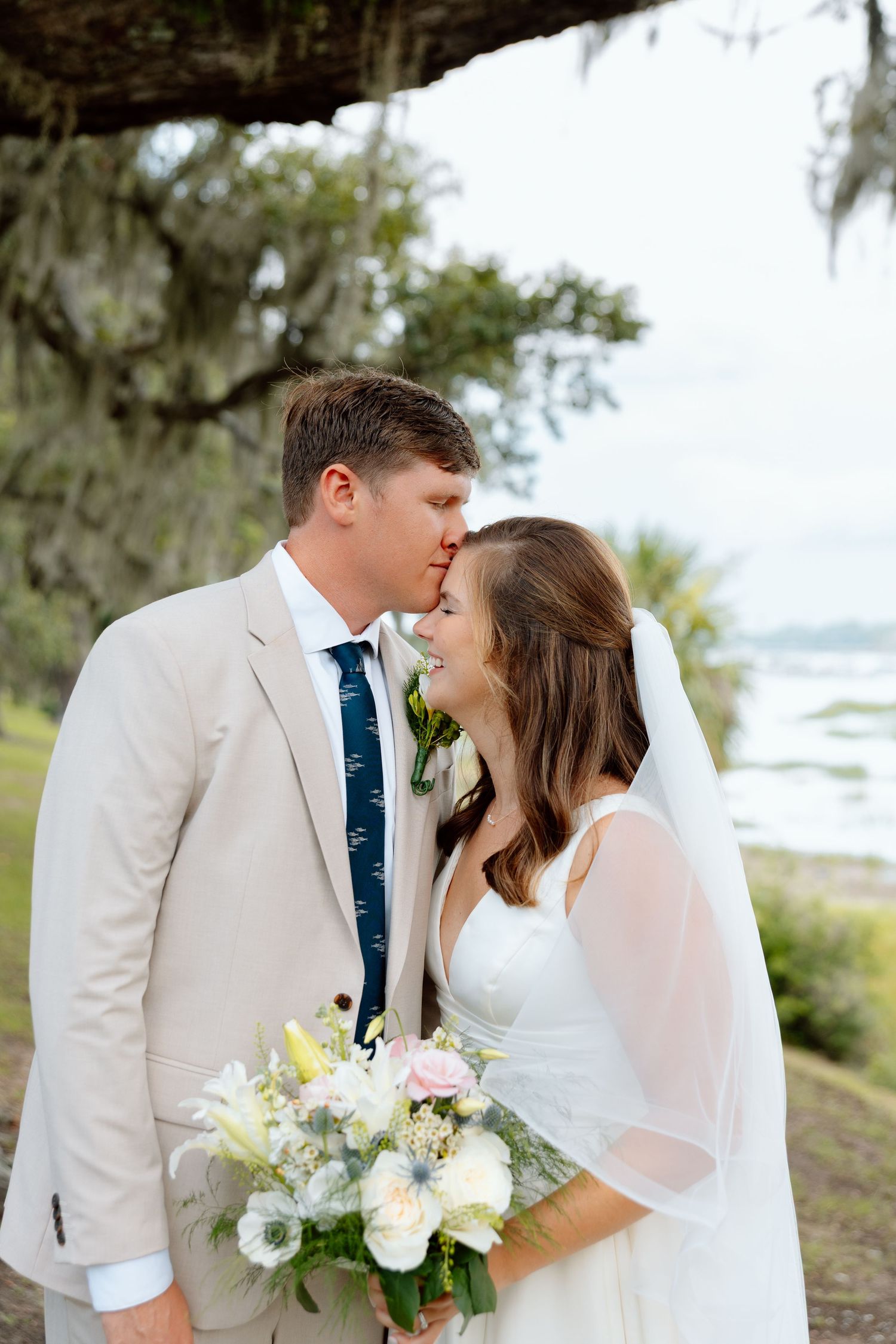 Bride and groom share a romantic moment under Spanish moss by a coastal marsh on their wedding day.
