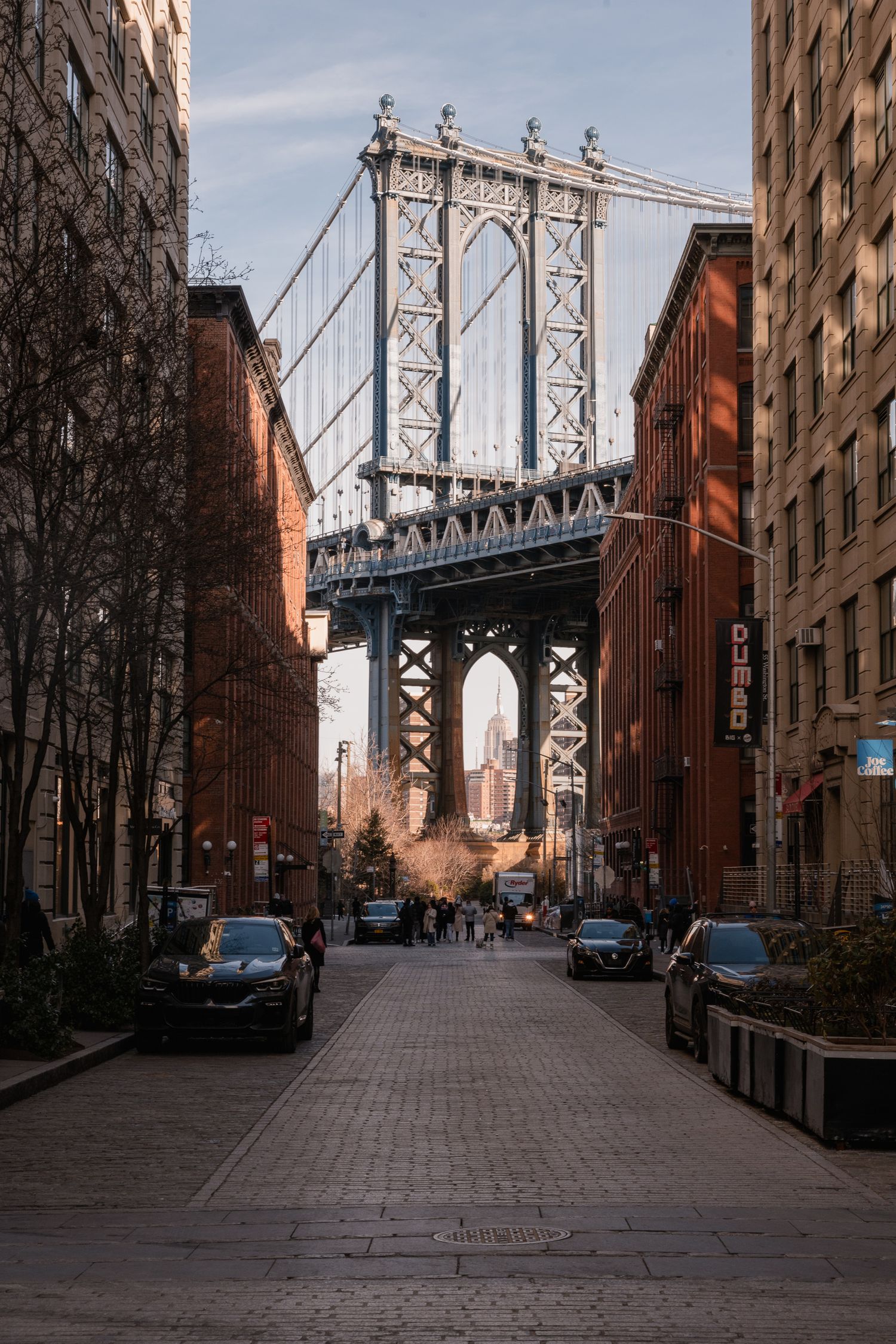 Vista iconica del Manhattan Bridge incorniciato tra i palazzi in mattoni rossi di Dumbo a New York, con auto e persone sulla strada in ciott