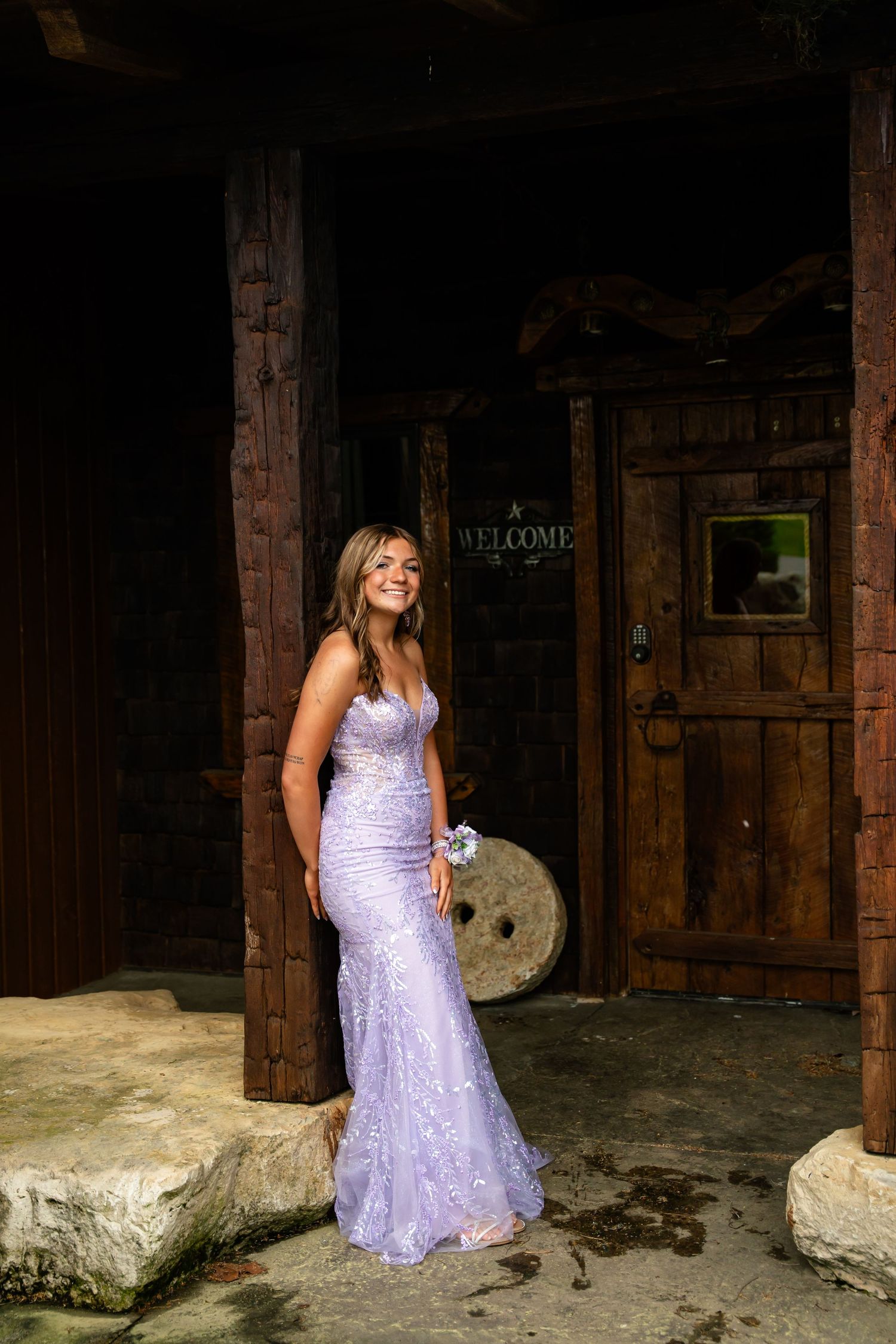 Young woman in lavender sequin mermaid prom dress with wrist corsage posing at rustic wooden entrance.