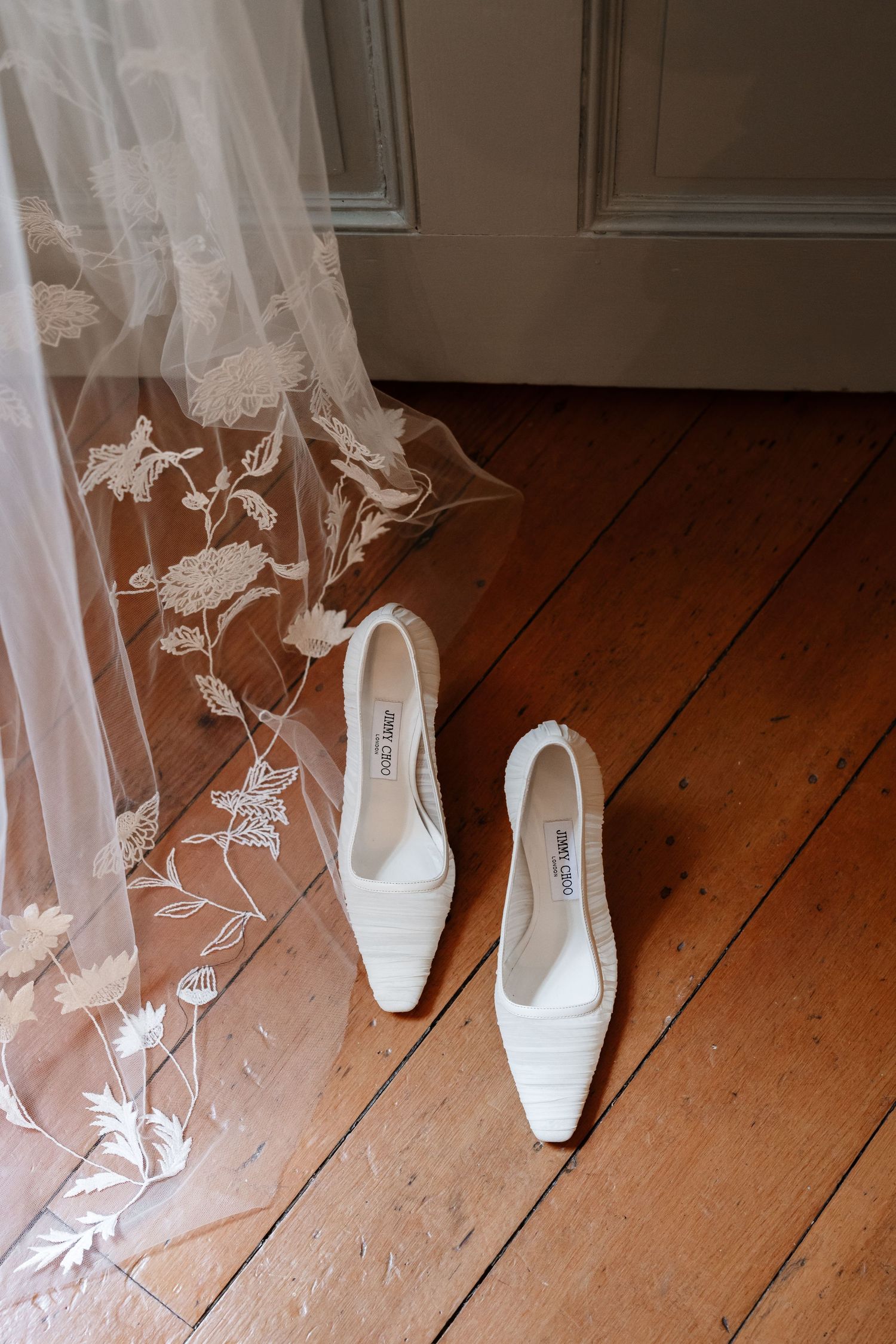 White Jimmy Choo bridal heels rest on hardwood floor beside an embroidered floral lace wedding veil.