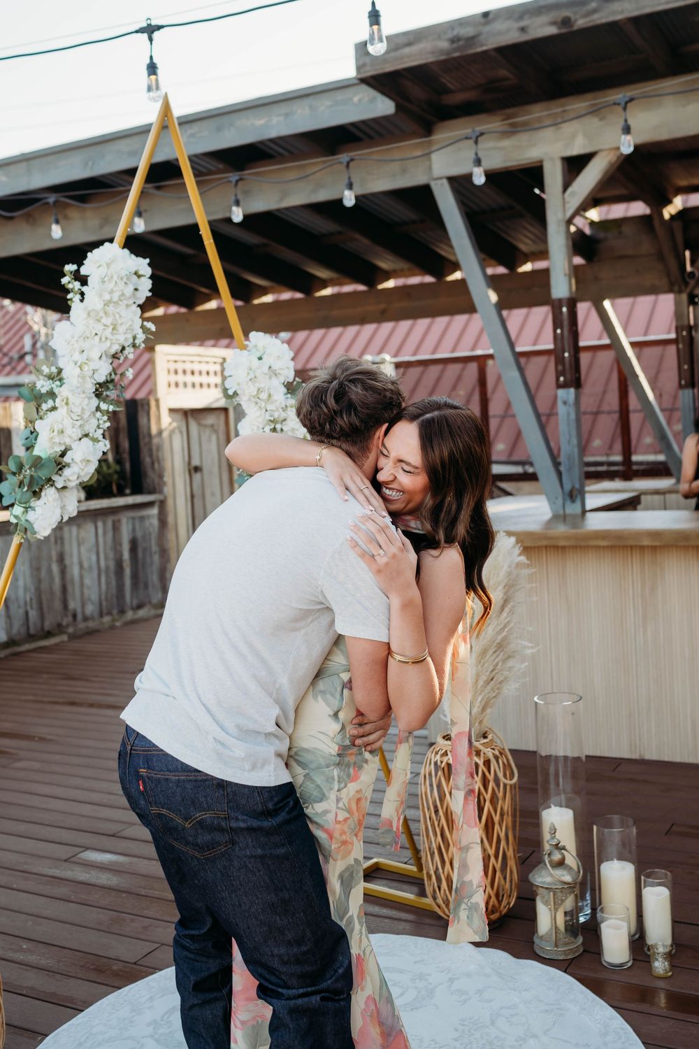 Couple sharing a joyful hug on a decorated rooftop venue with white flowers, candles, and string lights at dusk.