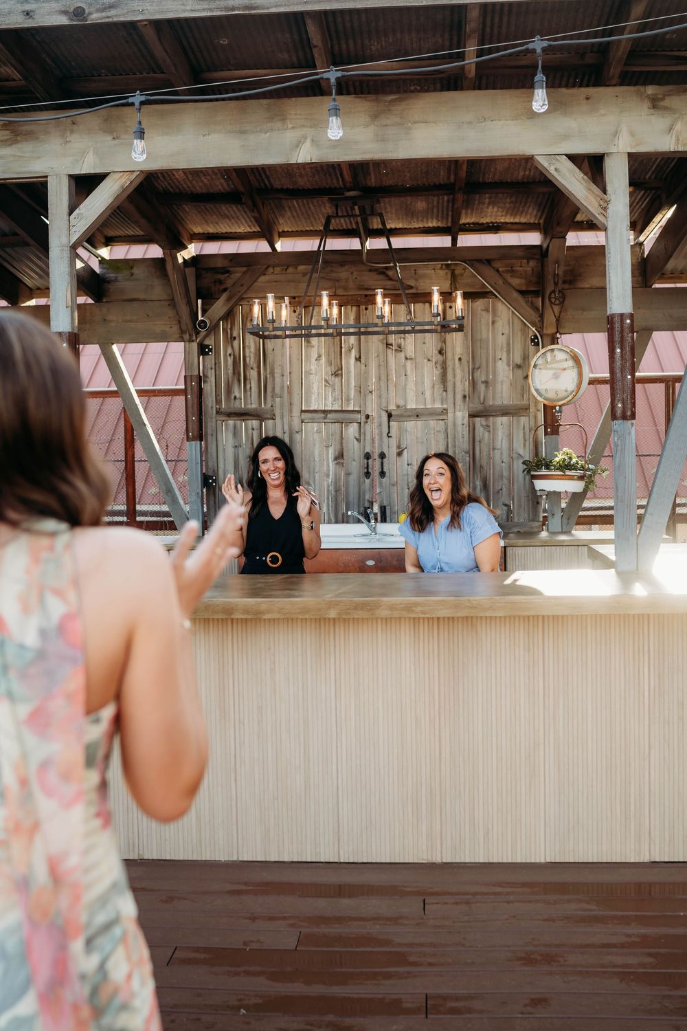 Two women laughing behind a rustic wooden bar counter in an open-air barn venue with chandelier lighting.