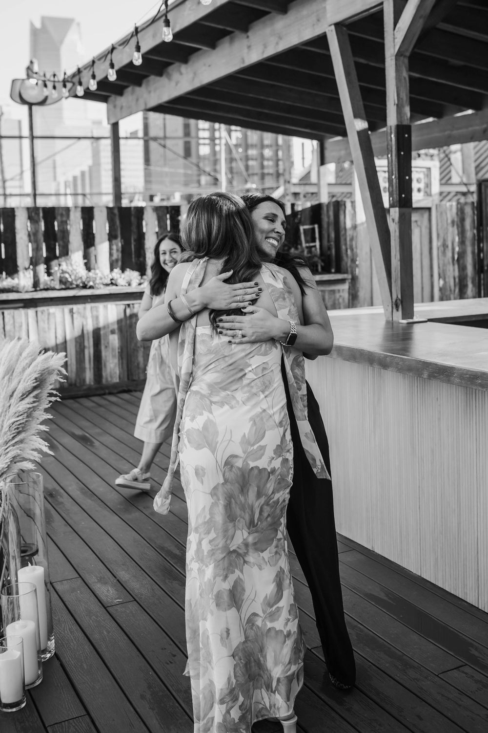 Black and white photo of two women sharing a warm hug on a rooftop venue deck during a wedding celebration.