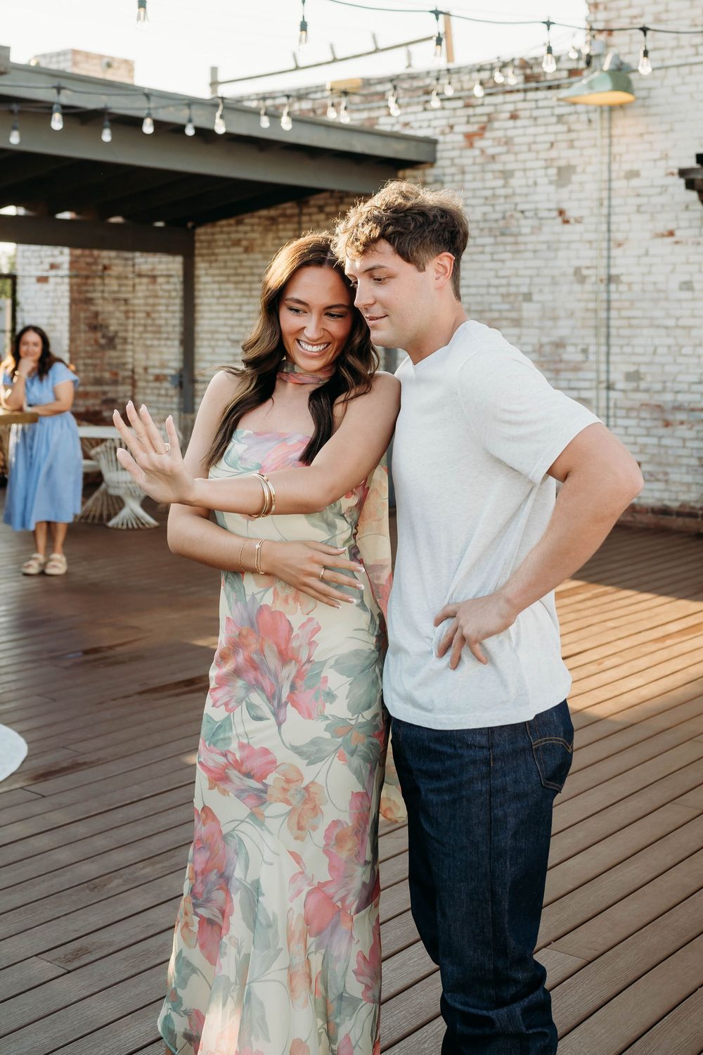 Happy couple on rooftop terrace, woman in floral dress showing engagement ring, man in white tee embracing her.