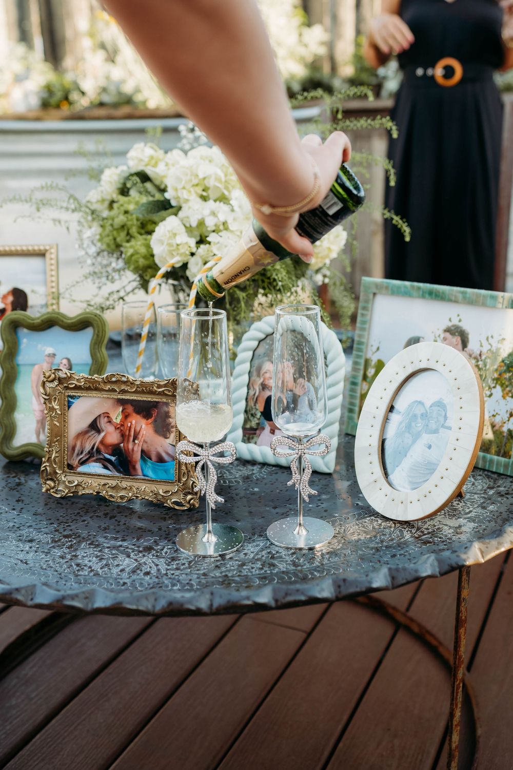 Champagne being poured into elegant glasses on a table decorated with framed photos and white flowers at a wedding reception.