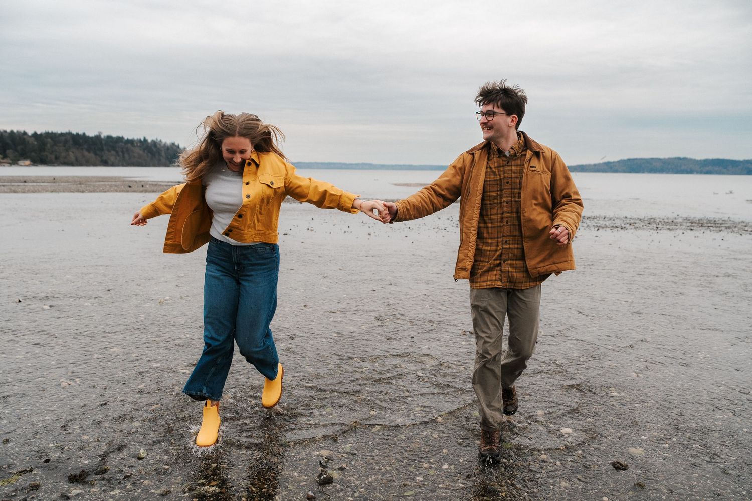 Couple holding hands while running on a wet beach wearing yellow rain boots and fall jackets for their engagement session in Seattle.