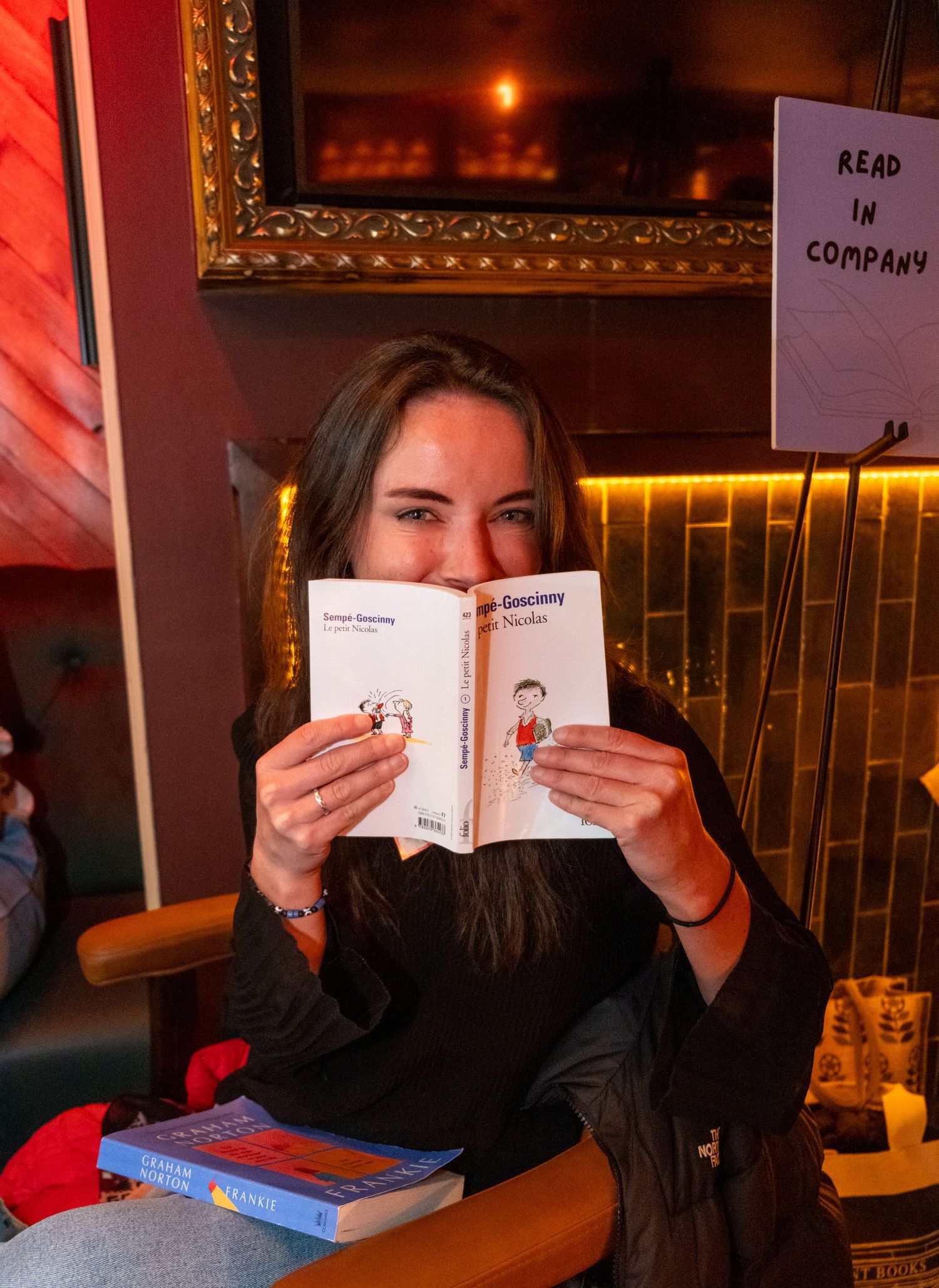 Woman smiling behind an open book at a cozy Read in Company event, with stacked books on the table.