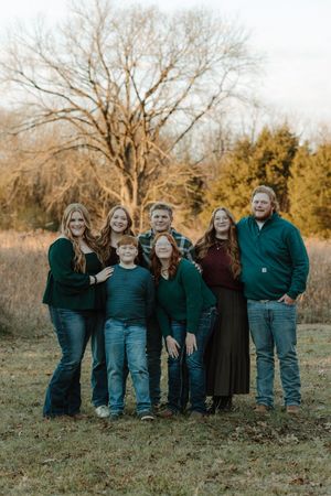 Family of seven posing outdoors in green and teal outfits among autumn trees and golden grass.