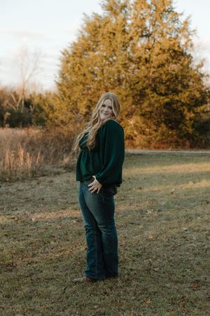 Young blonde woman in green top and jeans smiling outdoors in a golden autumn field with trees.
