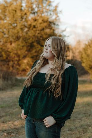 Young woman with long wavy blonde hair wearing a dark green off-shoulder top and jeans, posing outdoors in autumn.