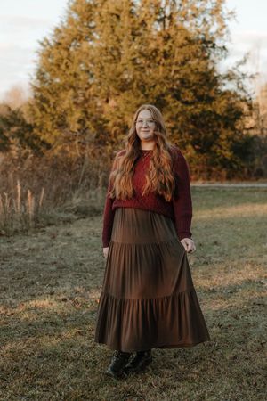 Young woman with long red hair wearing a burgundy sweater and brown tiered maxi skirt standing in a fall outdoor setting.