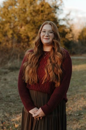 Young woman with long wavy red hair wearing a burgundy sweater and olive skirt, smiling outdoors in autumn setting.