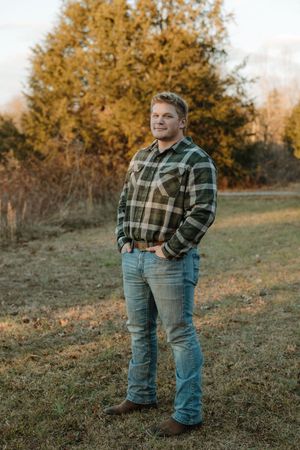 Young man in plaid flannel shirt and jeans standing in autumn field smiling.