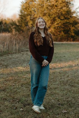 Young woman in burgundy sweater and jeans posing outdoors in fall setting.