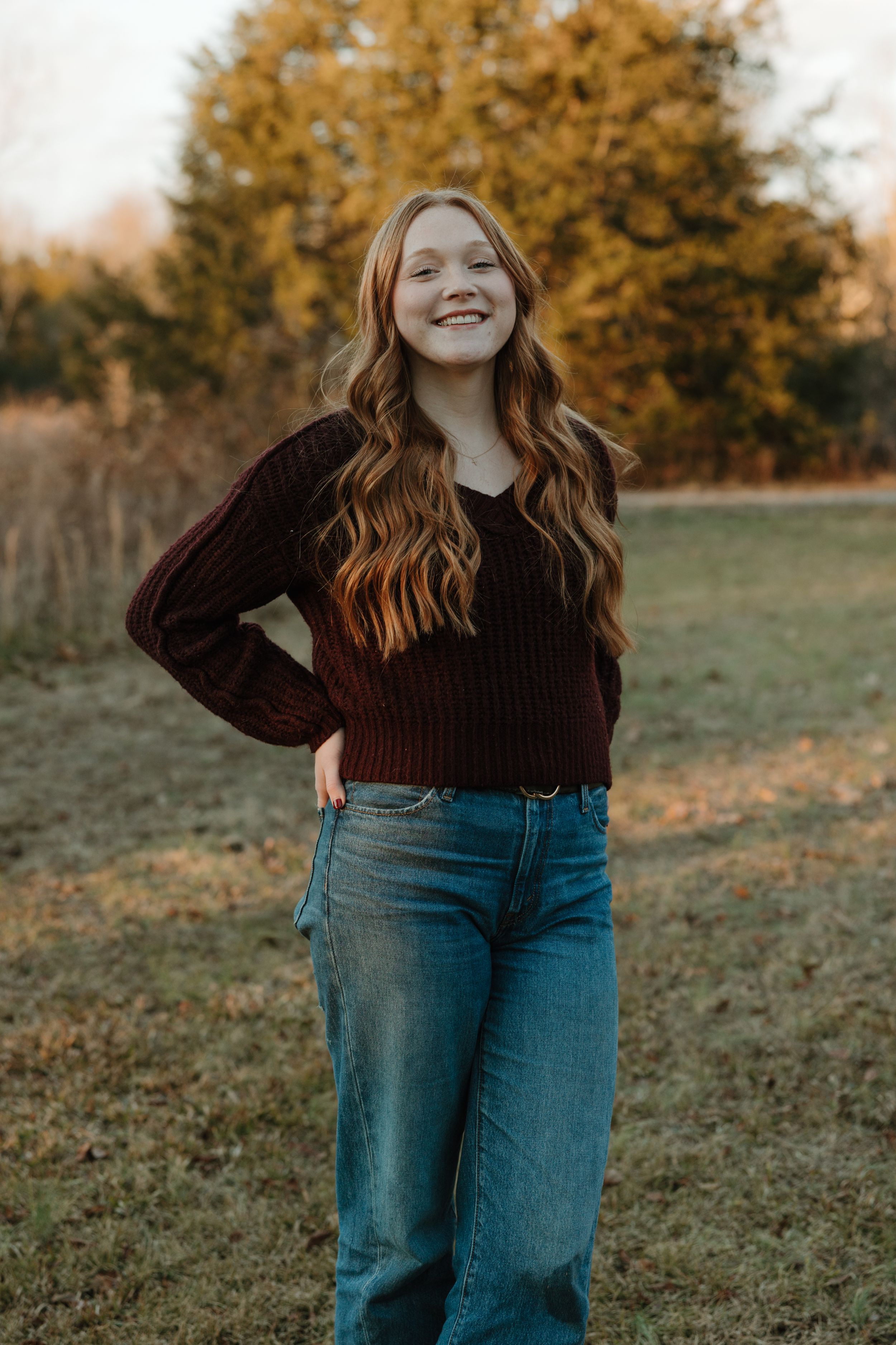 Young woman with long wavy hair smiling outdoors in a dark red sweater and jeans during autumn.
