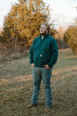 Man in green Carhartt pullover and jeans stands in autumn field with golden trees in background.