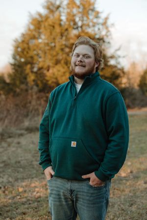 Young man in green Carhartt pullover smiling outdoors among autumn trees in a field.
