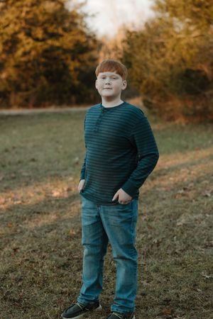 Red-haired boy in striped teal shirt and jeans standing in a grassy field with autumn trees in background.