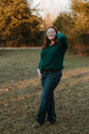 Young woman in green sweater and jeans poses outdoors in a field with autumn trees in the background.