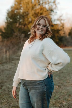 Young woman in cream waffle knit sweater and jeans smiling outdoors during golden hour.