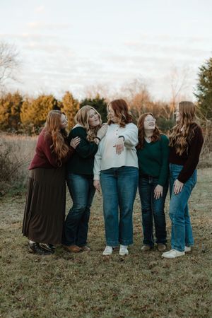 Five women laughing and posing together outdoors in a field during golden hour, wearing casual fall outfits.
