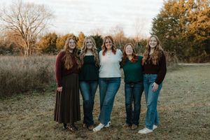 Five women smiling together outdoors in a fall field, wearing dark green, white, and burgundy outfits.