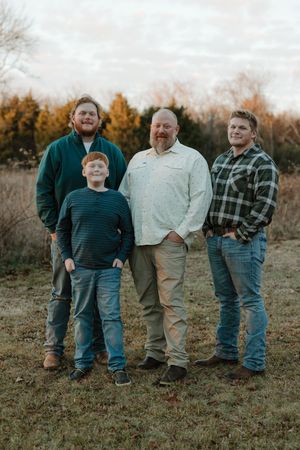 Father poses with three sons in casual attire in an outdoor field during autumn sunset.