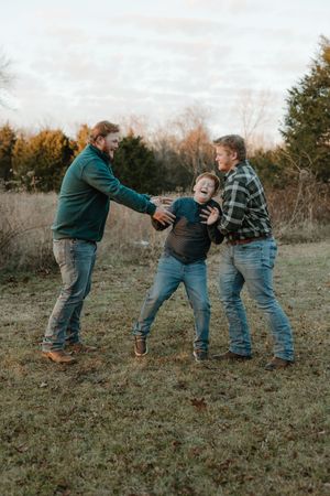 Two men playfully tickle a laughing boy outdoors in a grassy field with trees in the background.