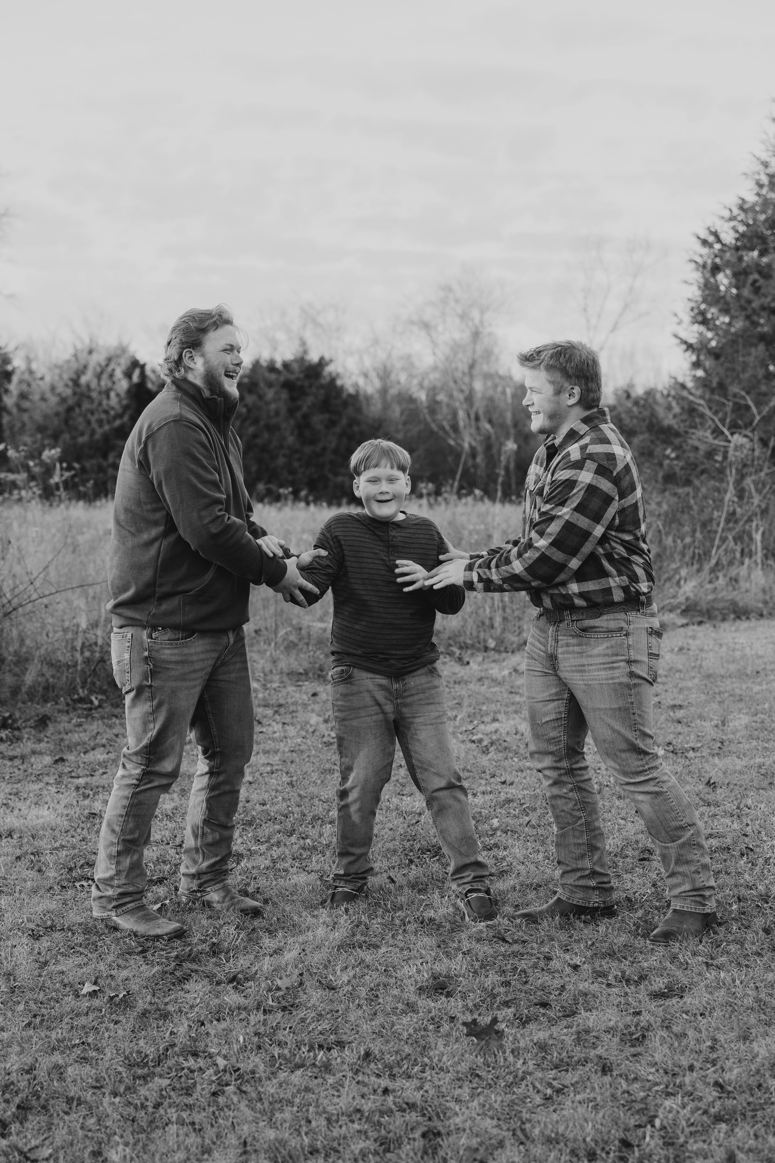 Black and white photo of two men and a young boy laughing together outdoors in a field with trees in the background.