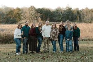 Large family of ten posing together in a field during autumn, wearing coordinated green, burgundy, and neutral tones.