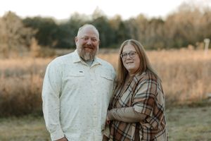 A smiling couple stands together in a golden field at sunset, man in white shirt and woman in plaid shawl.
