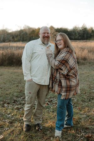 Smiling couple posing outdoors in a field during autumn, woman wearing plaid shawl and glasses, man in white shirt.