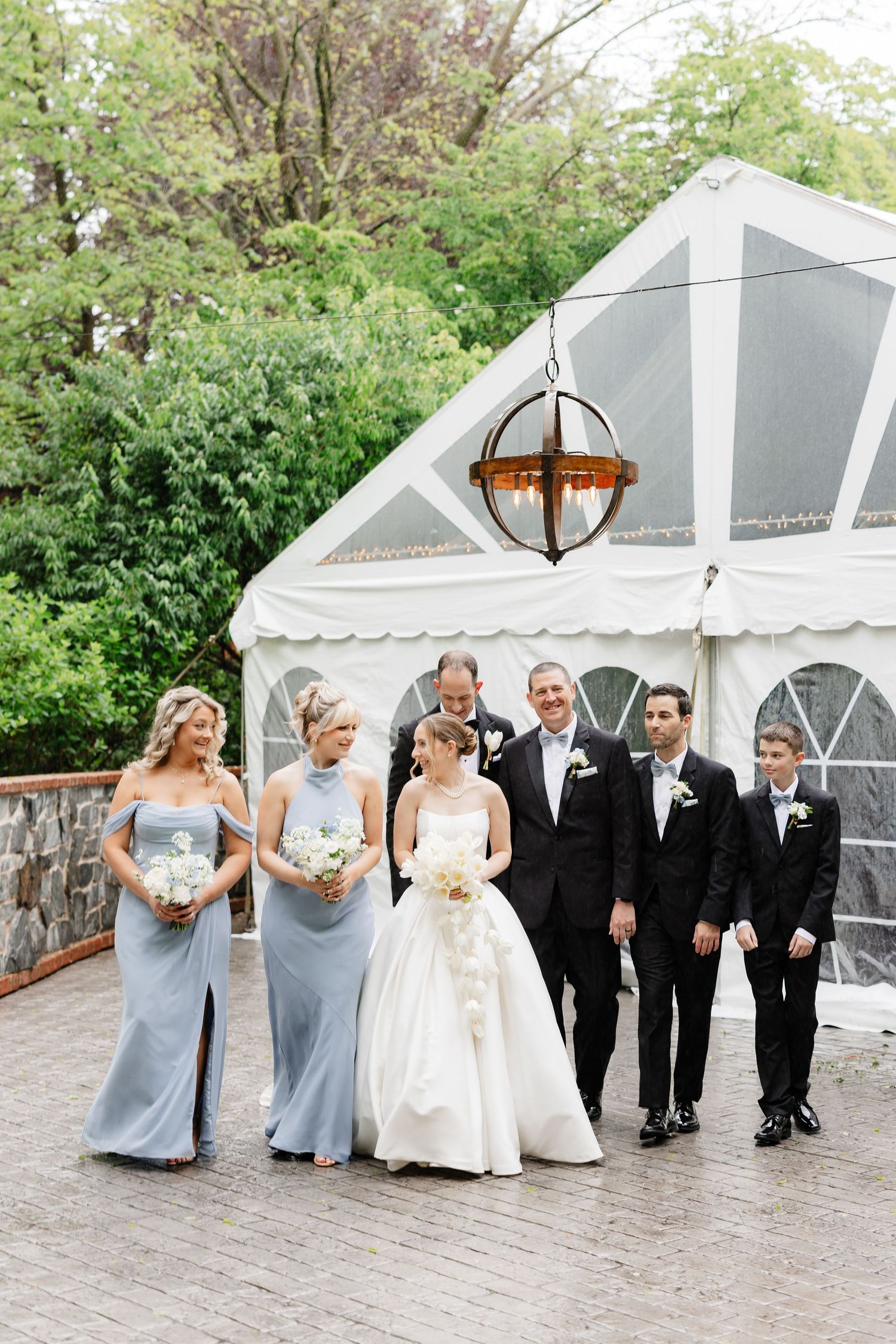 Wedding party walking outdoors near white tent venue, bridesmaids in dusty blue gowns and groomsmen in black tuxedos.